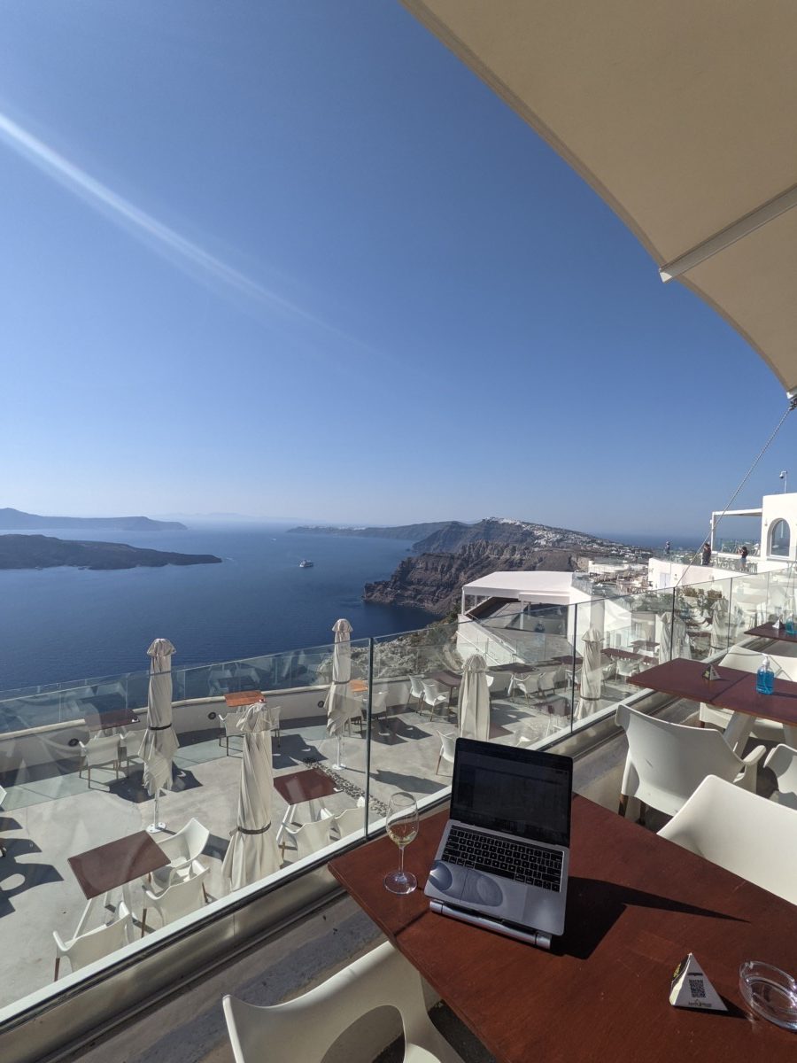 A laptop computer sits on a table with an empty glass of wine next to it at a winery in Greece that overlooks the ocean and a large patio with empty tables and chairs