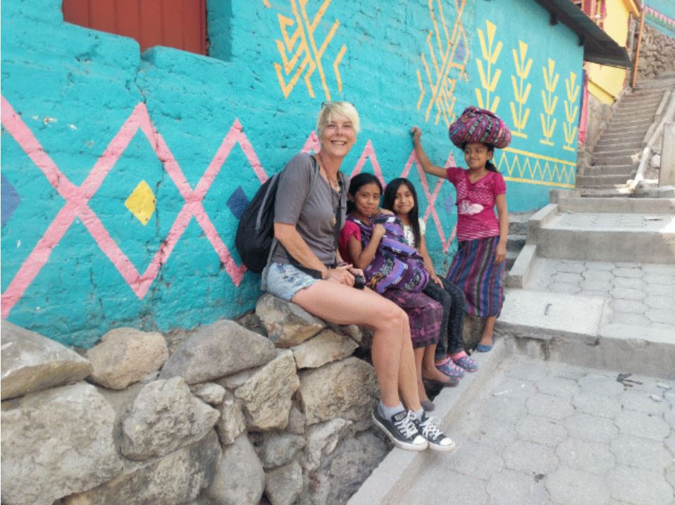 Candy sits on a low stone wall next to a turquoise painted building with three kids sitting next to her
