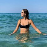 Chelsea Kane stands in waist-deep water in the ocean while looking off to the left with both arms out at her sides to touch the surface of the water