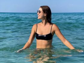 Chelsea Kane stands in waist-deep water in the ocean while looking off to the left with both arms out at her sides to touch the surface of the water