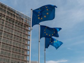 Three European Union flags fly in the wind in front of a large and tall building in Brussels