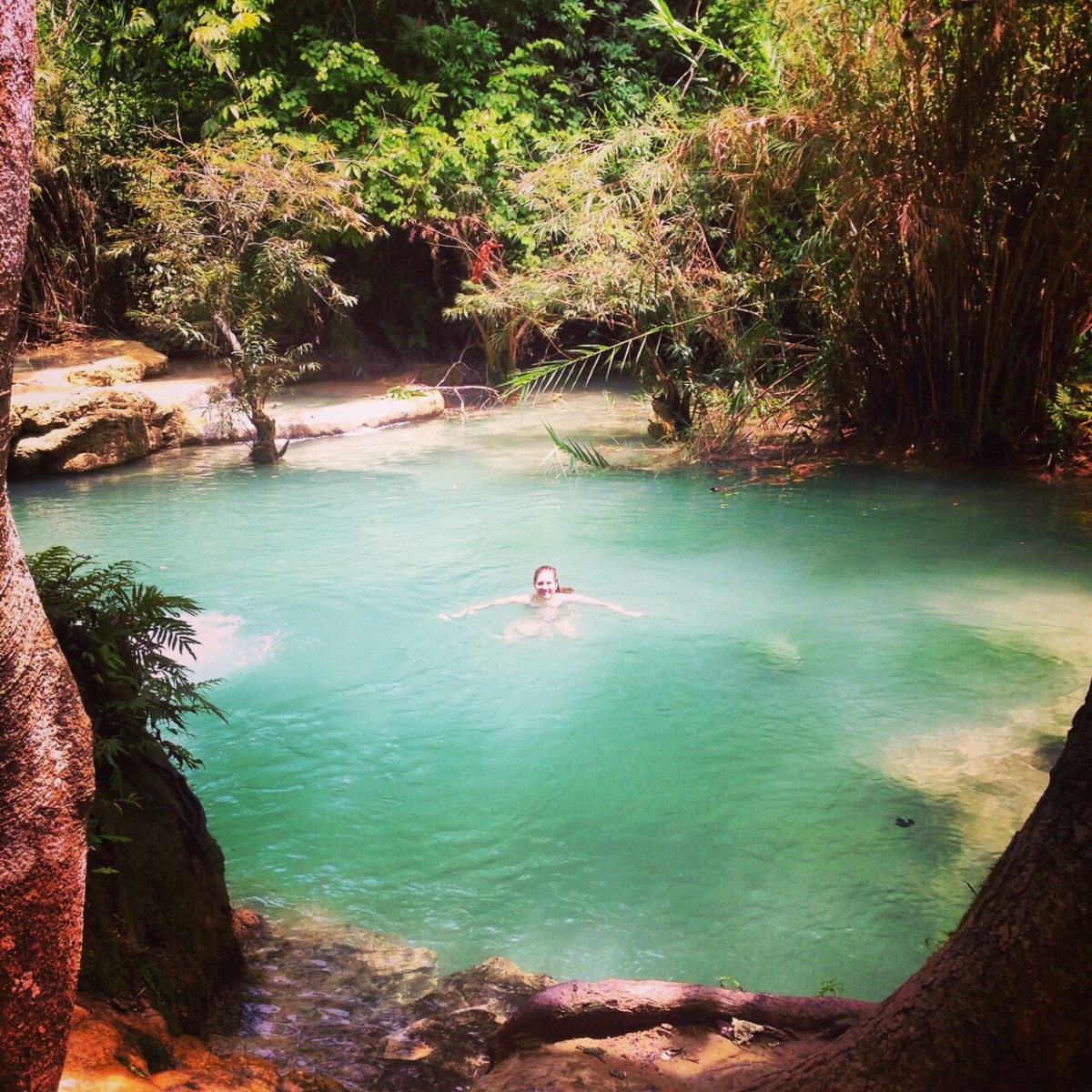 Katherine Conaway swims in the middle of an emerald green pool in Laos with green trees around the swimming hole