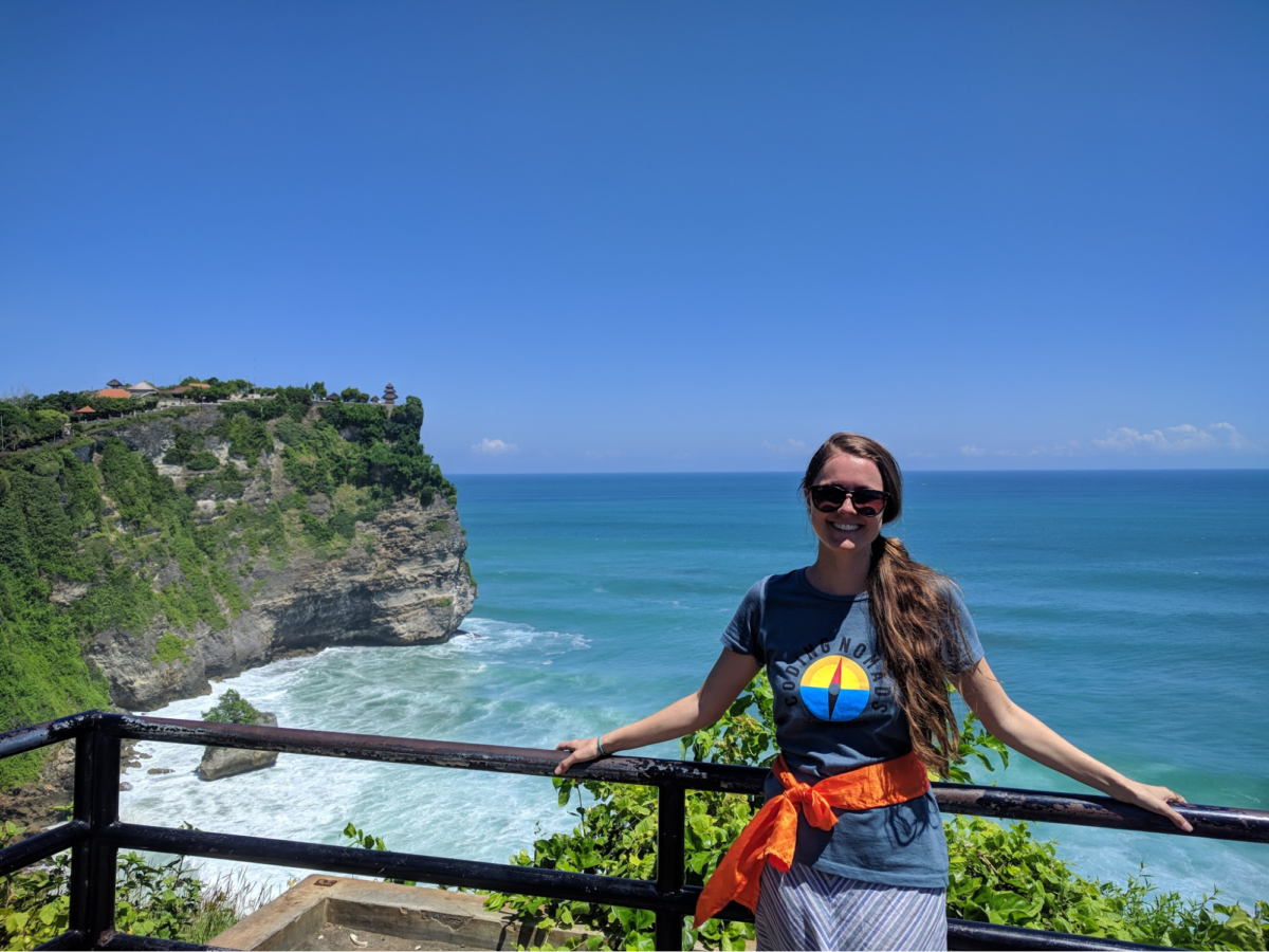 a woman stands with both arms and hands on a wooden railing on a cliff that overlooks the ocean 