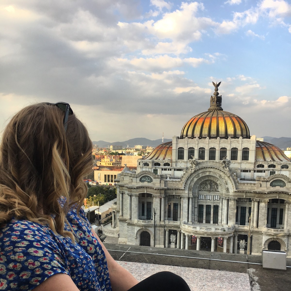Katherine Conaway sits outside on Mexico city and looks into a large old building that has the sun shining on the domed roof to make it green, yellow, and orange