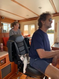 Katherine Conaways father sits driving their boat with a cat sleeping on the chair behind him while Katherine's mom looks on from the back of the cabin