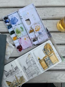 Watercolor paints sit next to three sketched drawings of a building with one partially painted in