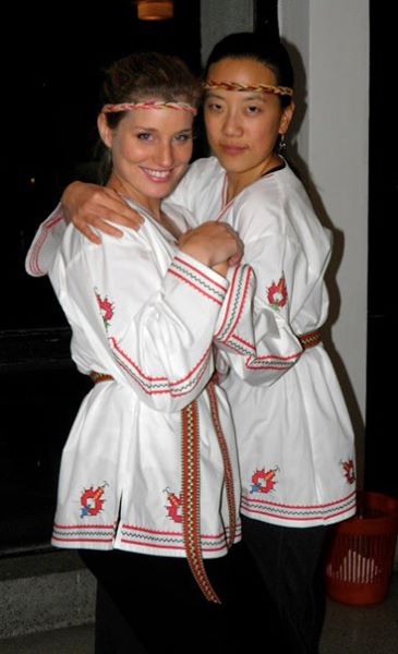 Katherine Conaway stands facing a friend while both are wearing white robes with red embroidery and matching headbands
