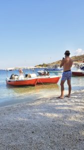 Dirk Bruwer stands facing two identical red and white boats in the water in Greece as he stands on a sandy shore in the shade