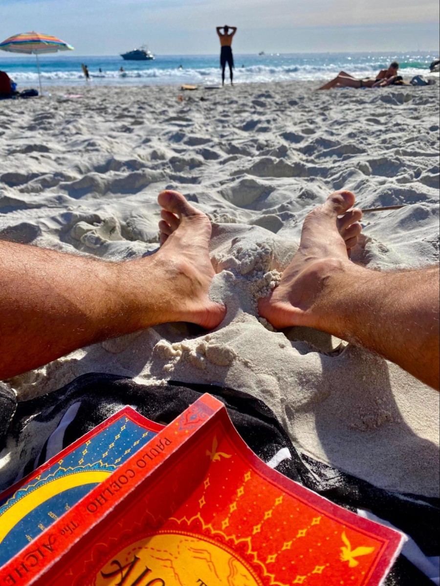 the book the alchemist sits open and face down on a towel on a sandy beach between a person's legs with the ocean nearby