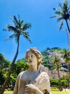 A white statue of a female sits between palm trees with greenery and a mountain behind the statue