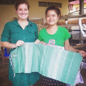 Katherine Conaway wears a green dress and stands next to a woman wearing a green shirt while both hold up a green tapestry that Katherine wove with the help of the other woman