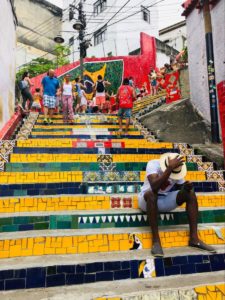 a colorful staircase in Rio with one man sitting on the steps and many others walking on the stairs
