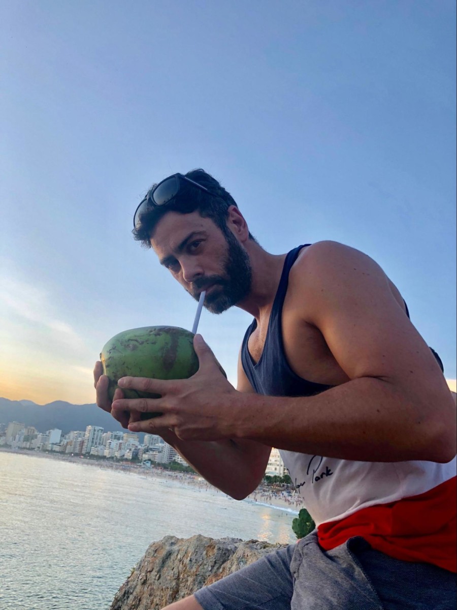 Dirk Bruwer stands next to the ocean while holding a large green coconut in both hands and he sips the coconut juice from a straw protruding from the coconut