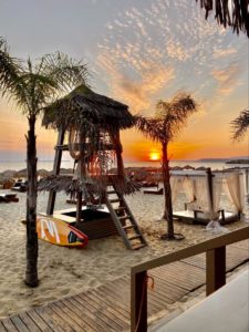 a tall lifeguard tower with a palm frond roof sits in a sandy beach at sunset in Greece with the ocean in the background