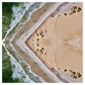 A photo taken from a drone shows an aerial view of the ocean with a sandy shore in a triangle on the right and crashing waves on the left