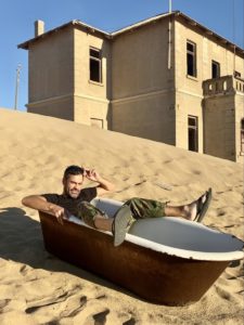 Dirk sits in an empty bathtub with one foot hanging over each side as the tub sits on a sand dune with a large house behind it