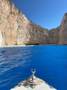 the bow of a boat in blue water in Greece with a large sand-colored cliff ahead on all sides of the boat