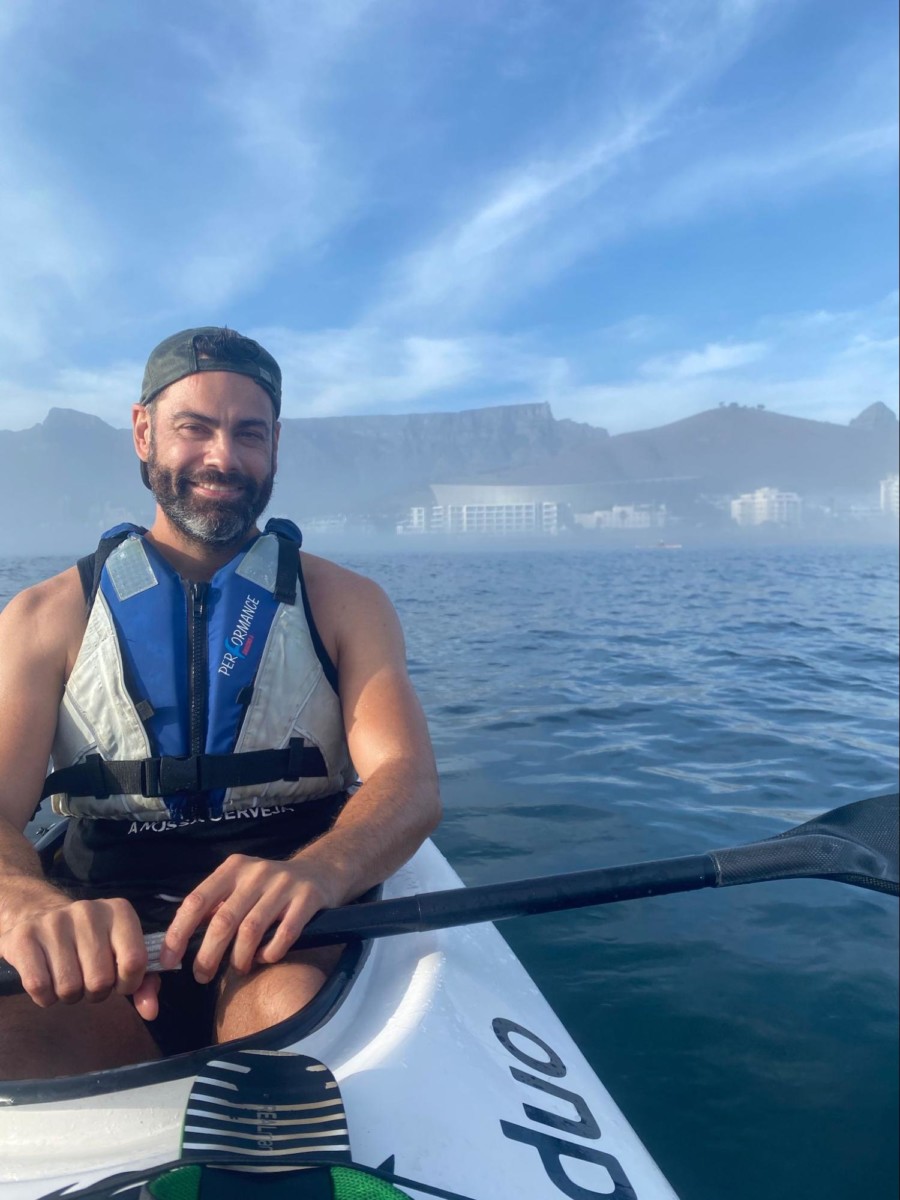 DIrk Bruwer sits in a white kayak on the ocean with the paddle sitting across the boat as he wears a life vest with cliffs in the background and some clouds in the blue sky 