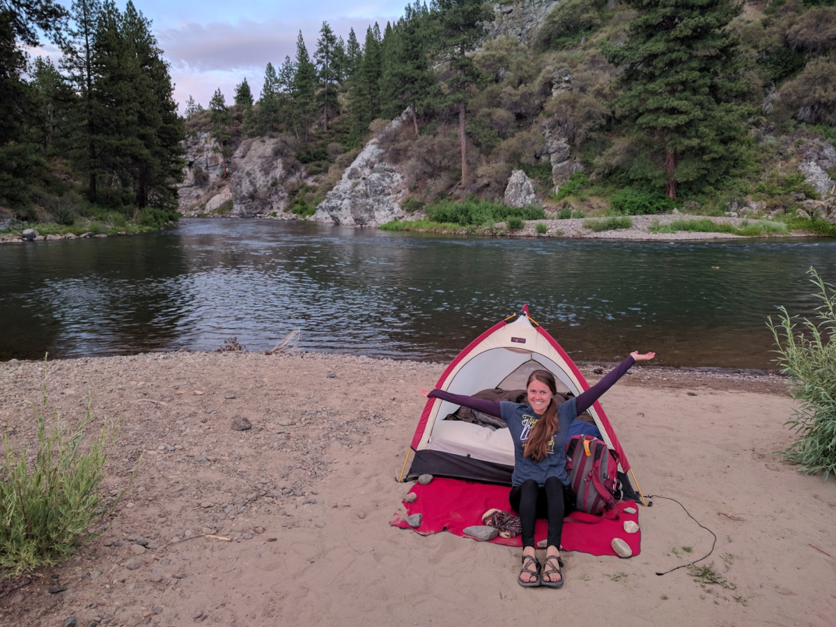 A woman sits in the entrance of a red and white tent with her hands in the air while the tent sits on a sandy area near a calm lake