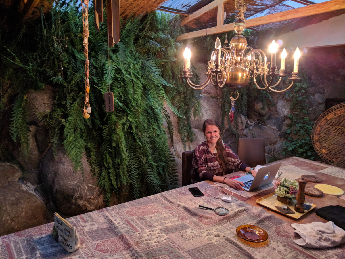 A woman sits at a table with a large chandelier above with her laptop open on the table in front of her 