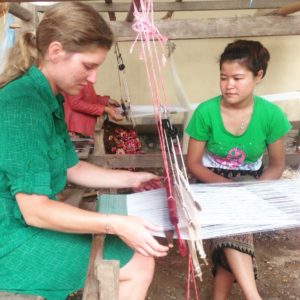 Katherine conaway sits in a green dress working on a loom in front of her while another woman wearing a green shirt looks at the loom