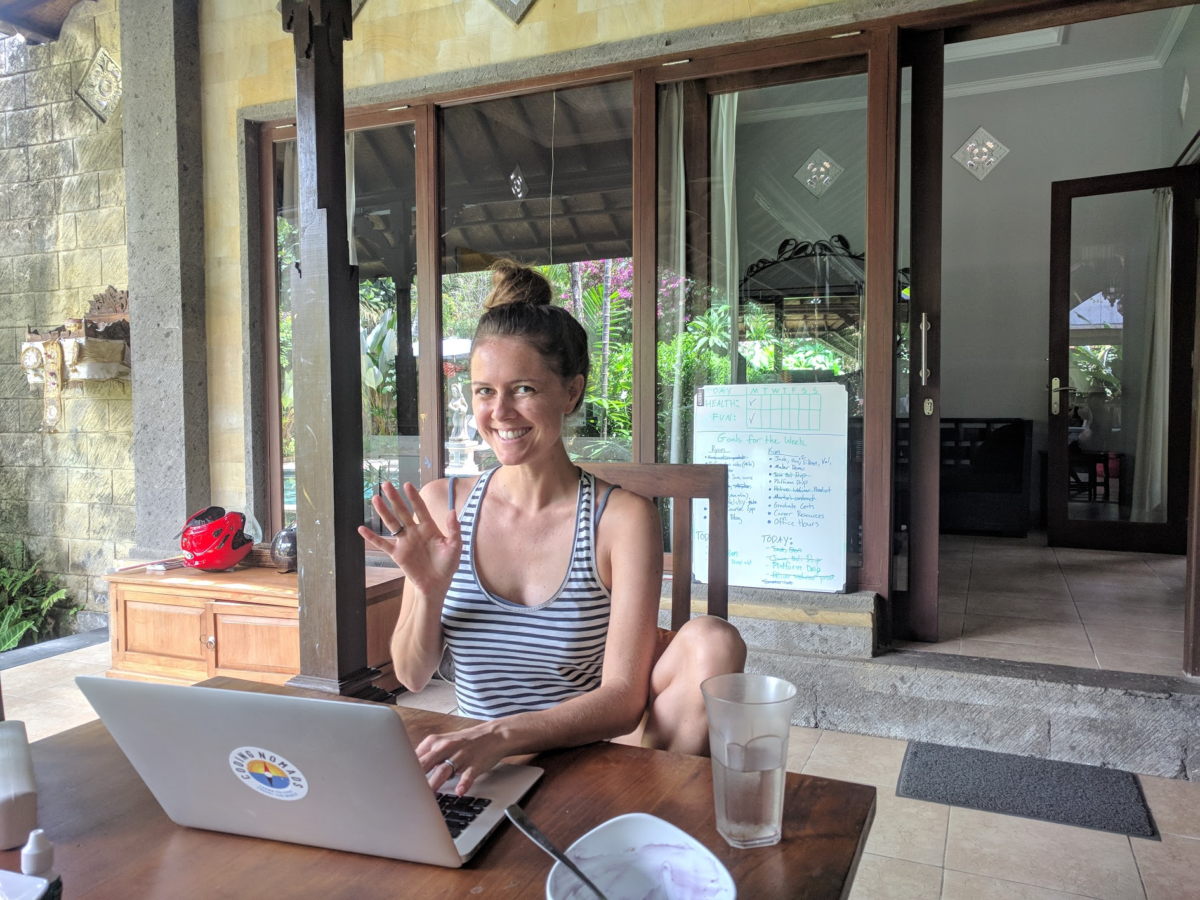 A woman waves while sitting at a table in front of her laptop at a table outside with a sliding glass door into the house behind her