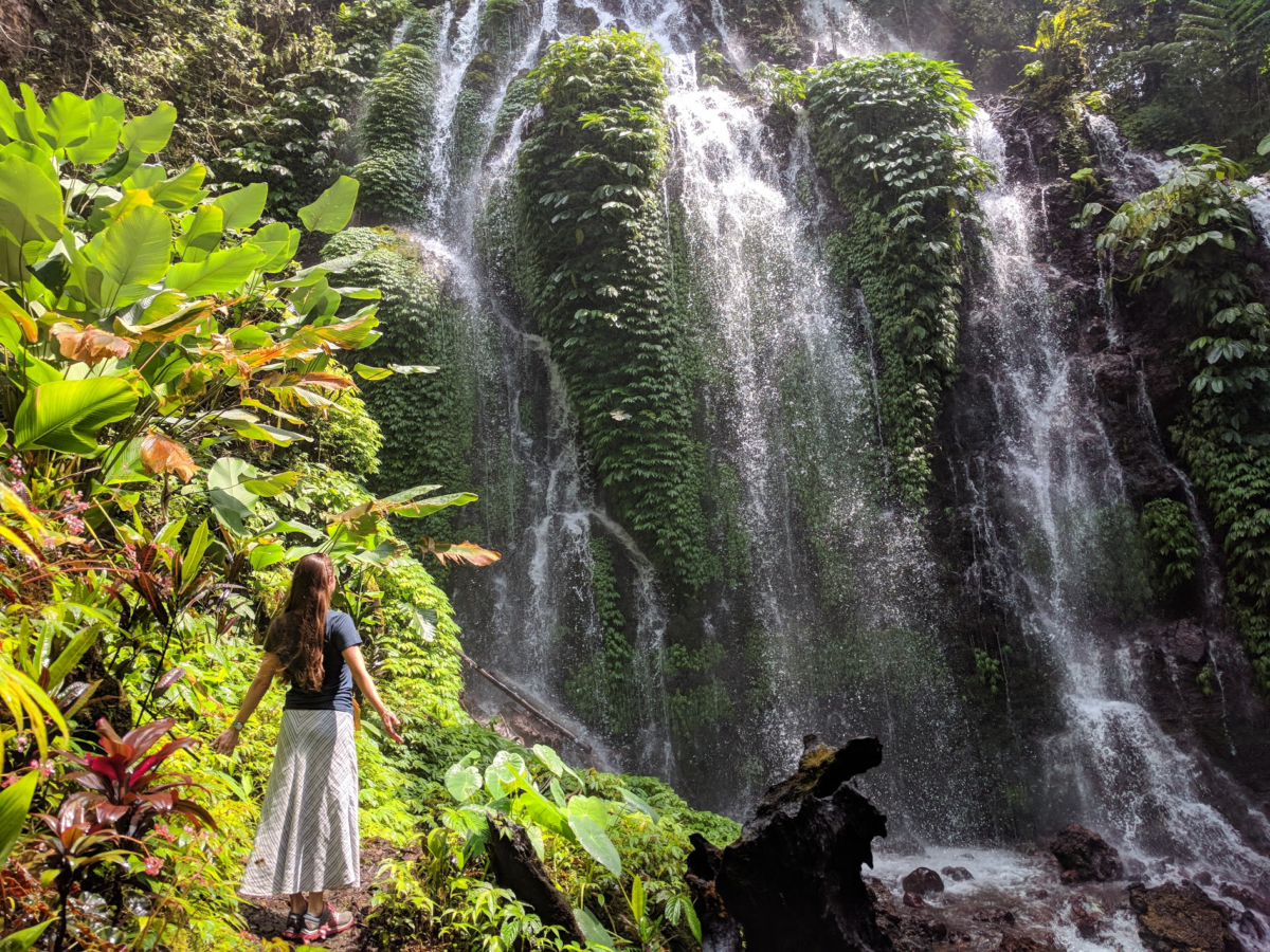 A woman stands with open arms facing a large waterfall that cascades down a lush rockface with greenery around