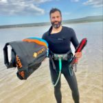 Dirk bruwer stands in ankle-deep water wearing a wetsuit and holding kiteboarding gear in both hands and under one arm