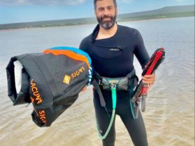 Dirk bruwer stands in ankle-deep water wearing a wetsuit and holding kiteboarding gear in both hands and under one arm