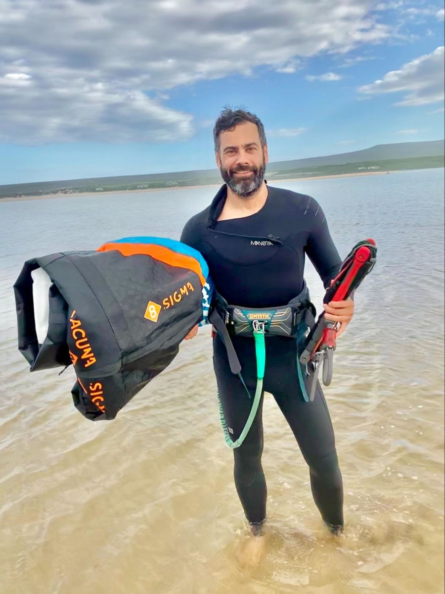 Dirk bruwer stands in ankle-deep water wearing a wetsuit and holding kiteboarding gear in both hands and under one arm