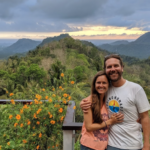 a male and female stand hugging sideways while standing in front of a wooden railing overlooking a valley and mountains with vegetation and flowers