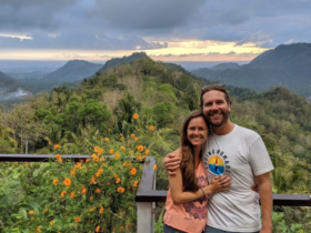 a male and female stand hugging sideways while standing in front of a wooden railing overlooking a valley and mountains with vegetation and flowers