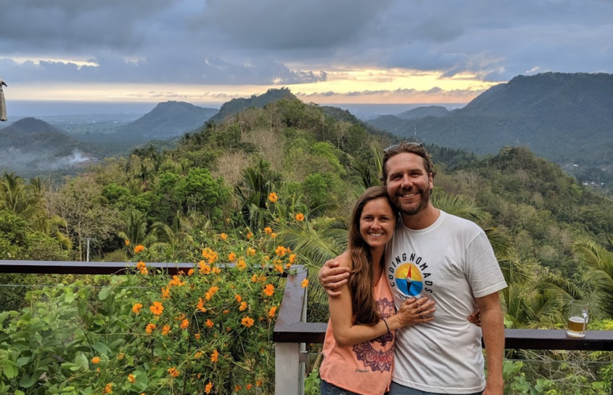 a male and female stand hugging sideways while standing in front of a wooden railing overlooking a valley and mountains with vegetation and flowers