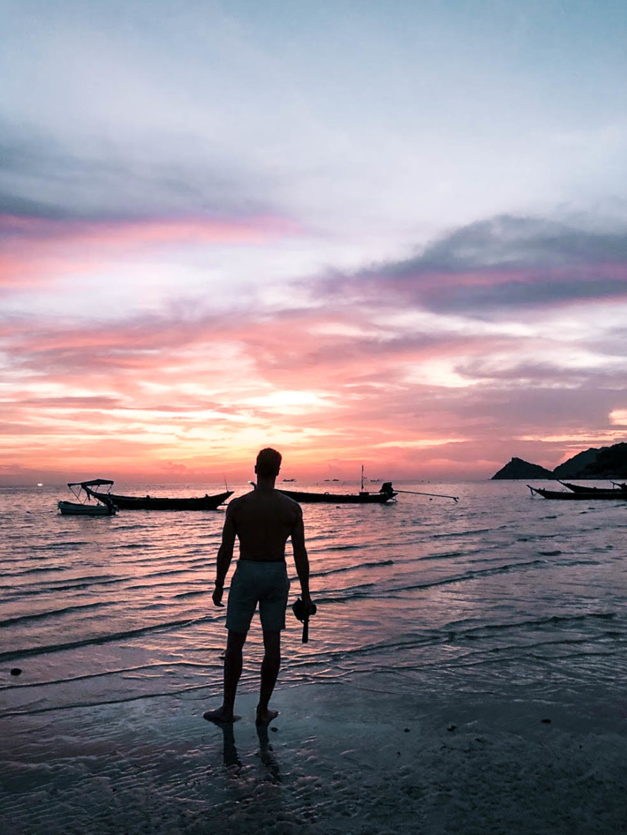 Blake Miner stands as a silhouette on a beach in Thailand during sunset with the sky purple, blue, orange, yellow and pink in front of him