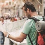 Caucasian guy carrying a red backpack with a yoga mat holder navigating an unfamiliar city with a map