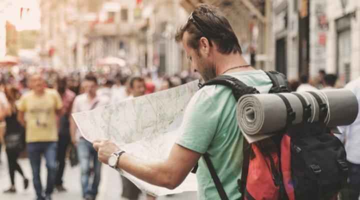 Caucasian guy carrying a red backpack with a yoga mat holder navigating an unfamiliar city with a map