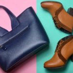 A travel garment bag on the floor beside a pair of brown ankle boots and a dark blue purse