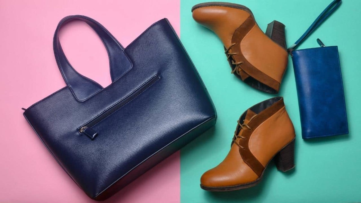 A travel garment bag on the floor beside a pair of brown ankle boots and a dark blue purse